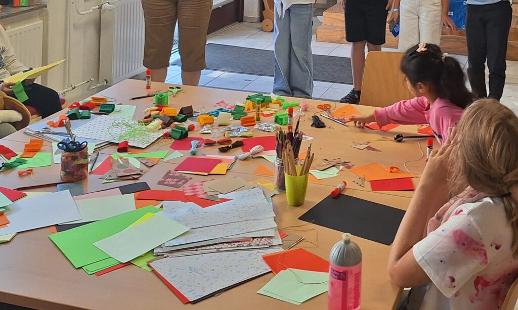 Children and adults stand and sit around a table covered with colorful paper, craft supplies, pencils, and glue, working on creative art projects together in a bright room.