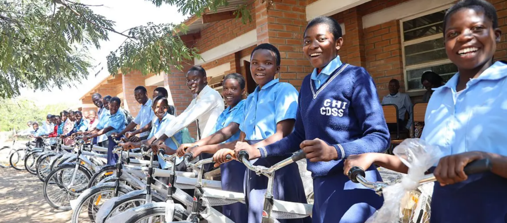 A group of smiling students in blue uniforms stand in a row outside with new bicycles, in front of a brick school building, enjoying a sunny day.