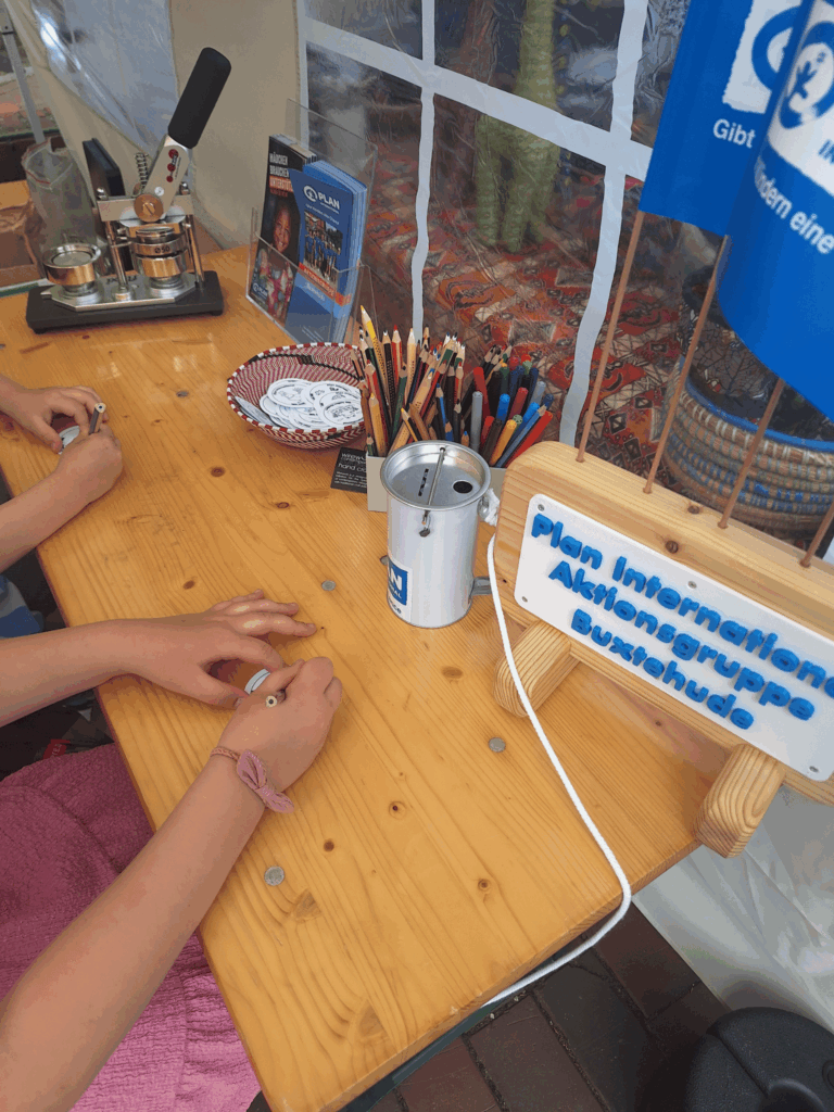 Two children sit at a wooden table with colored pencils, stickers, a donation can, and informational materials from Plan International. Signs and decorations are displayed, suggesting a charity or community event.