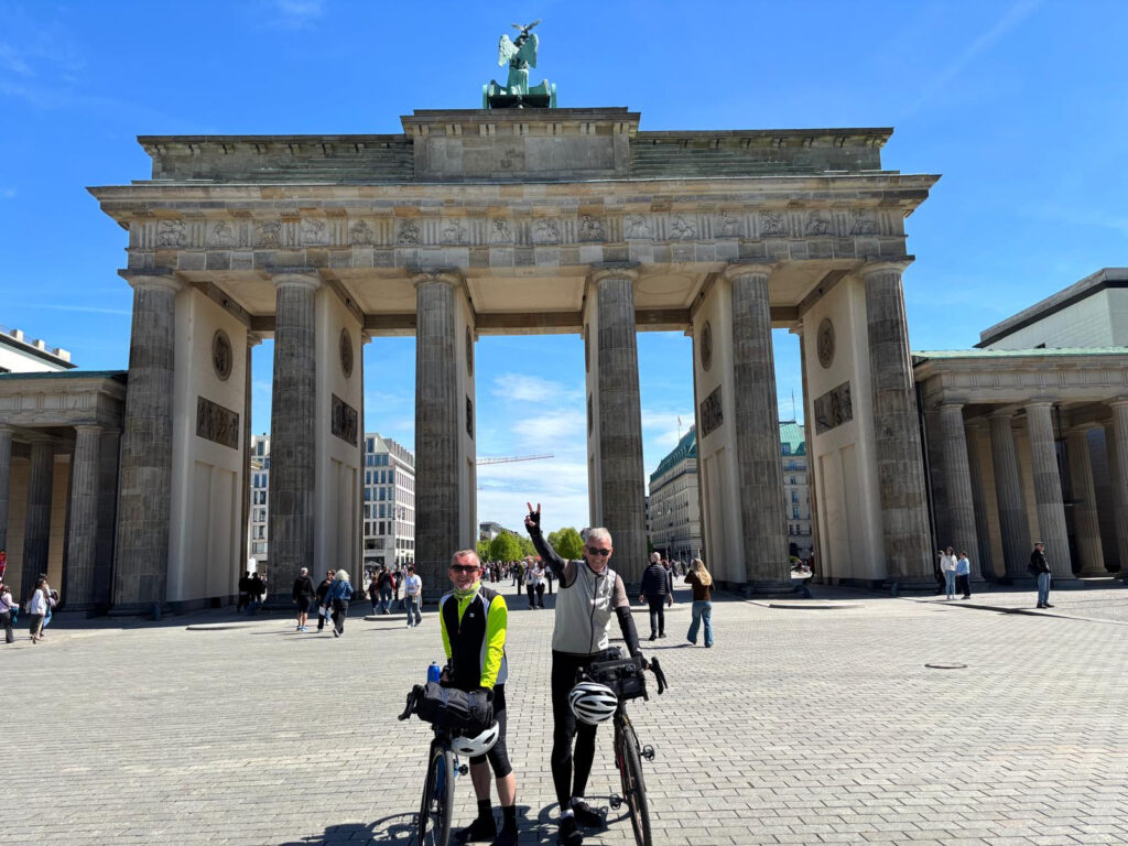 Zwei Radfahrer posieren an einem sonnigen Tag mit ihren Fahrrädern vor dem Brandenburger Tor in Berlin. Eine Person macht ein Friedenszeichen, während im Hintergrund Menschen spazieren gehen und sich versammeln.