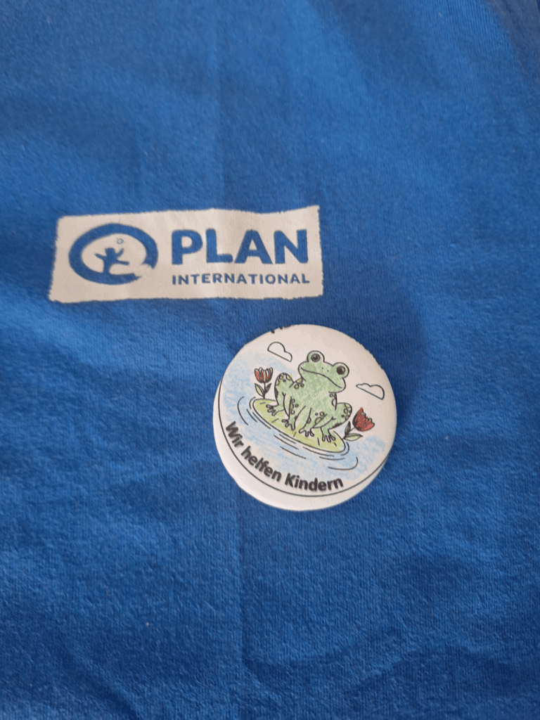 A blue fabric with a Plan International logo and a round badge featuring a green frog on a lily pad, water lilies, and the German text Wir helfen Kindern (We help children).
