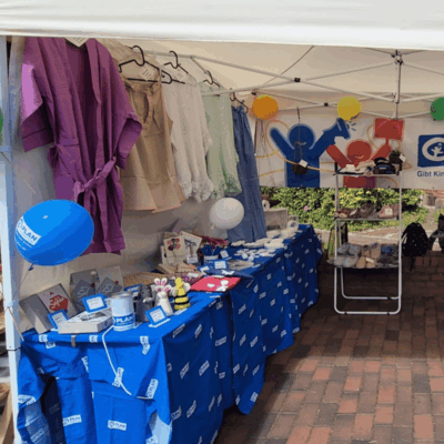A market stall under a white canopy displays clothing, balloons, toys, and informational materials on blue tablecloths. A banner with colorful heart and medical icons hangs at the back. Brick pavement and greenery are visible.