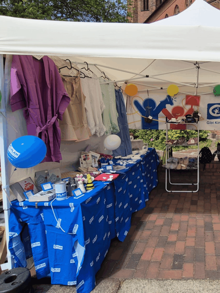 A market stall under a white canopy displays clothing, balloons, toys, and informational materials on blue tablecloths. A banner with colorful heart and medical icons hangs at the back. Brick pavement and greenery are visible.