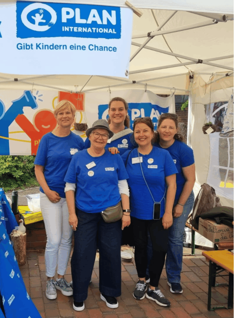 Five smiling women wearing matching blue Plan International shirts stand together under a booth tent with a banner reading Gibt Kindern eine Chance (Give children a chance in German).