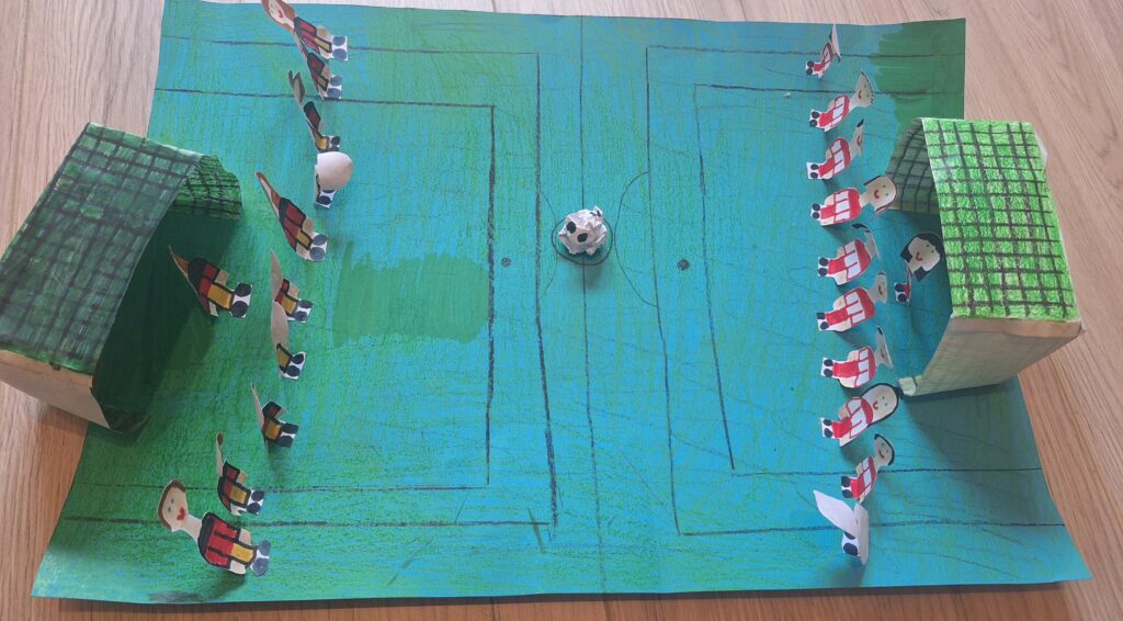Ein handgefertigtes Modell eines Fußballfeldes mit Papierspielern in roten oder schwarzen Trikots, zwei grünen Toren an jedem Ende und einem Papierfußball in der Mitte, alles auf einem blau-grünen Feld mit gezeichneten Linien.