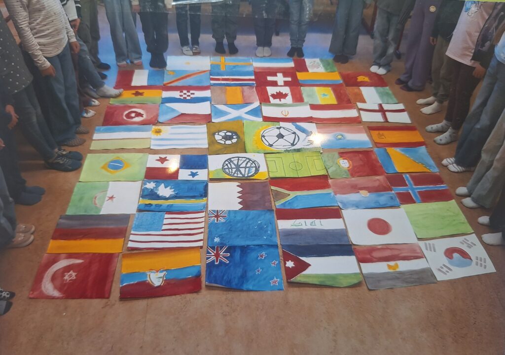 Die Kinder stehen in einem Kreis um gemalte Darstellungen verschiedener internationaler Flaggen, die auf dem Boden ausgelegt sind und die globale Einheit und Vielfalt symbolisieren.