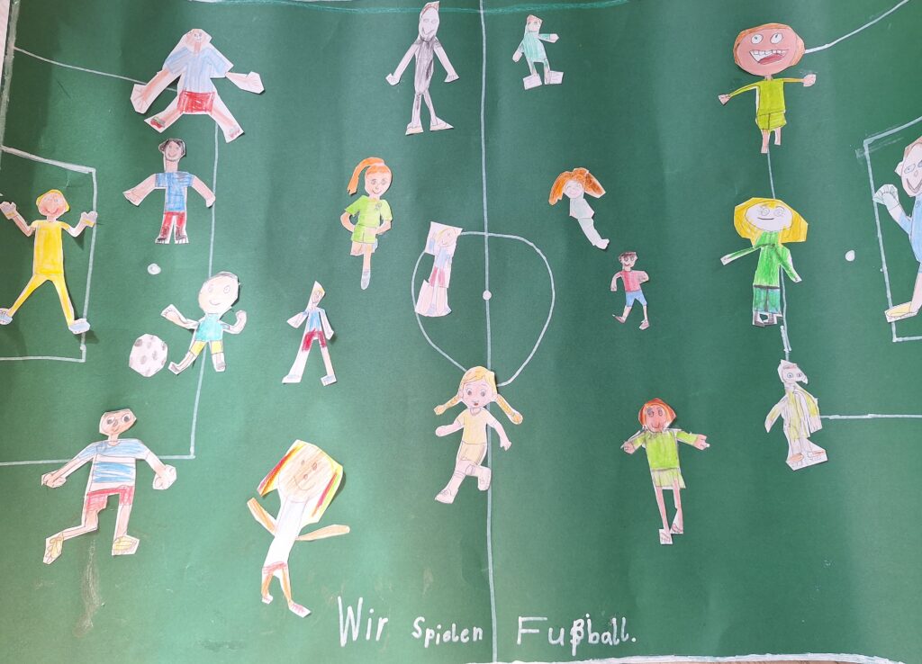 Kinderzeichnungen von Fußballspielern auf einer grünen Wiese, mit verschiedenen Figuren in verschiedenfarbigen Outfits, einem Fußball und dem Schriftzug Wir spielen Fußball am unteren Rand.